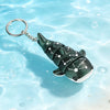 A dark green devil ray figurine with white star patterns floats in clear blue water attached to a silver keyring.