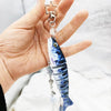 A hand holds a silver fish-themed acrylic keychain charm with a star top and a translucent fish pendant.