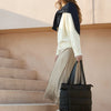 A woman wearing a cream sweater and beige skirt stands on stairs holding a black large capacity casual tote bag.