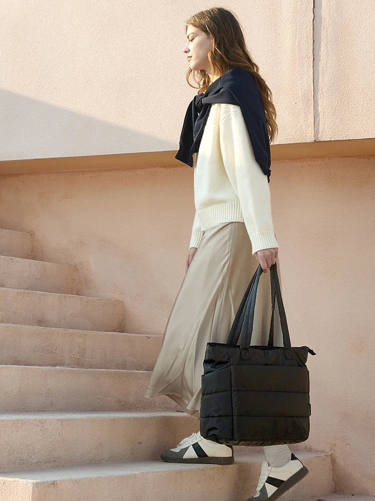 A woman wearing a cream sweater and beige skirt stands on stairs holding a black large capacity casual tote bag.