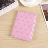 A pink heart print passport cover travel wallet resting on a light wooden surface next to a white cup and photo.