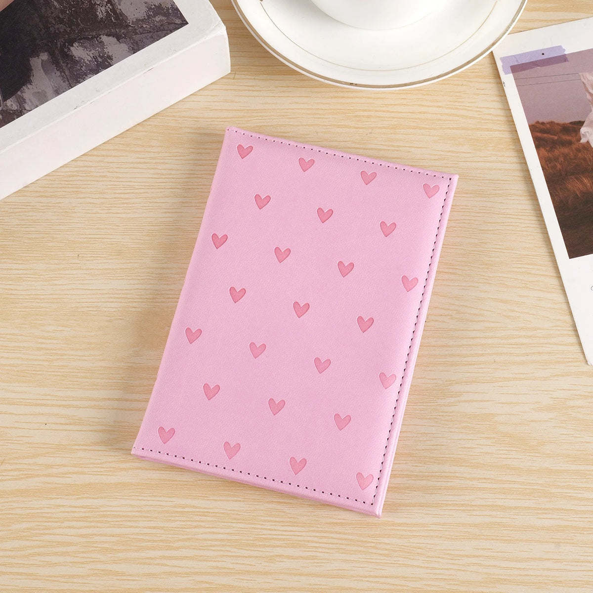 A pink heart print passport cover travel wallet resting on a light wooden surface next to a white cup and photo.