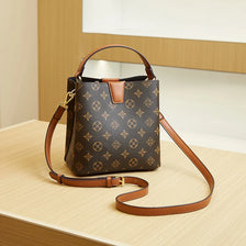 A brown leather bucket bag with a gold-toned strap and patterned fabric resting on a light wooden surface.