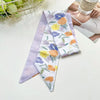 Une pile de foulards pour cheveux en soie élégants à motifs floraux violets, blancs et jaunes sur une surface blanche.