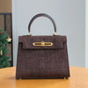 A dark brown crocodile pattern tote bag with a leather handle and gold hardware sitting on a wooden table.