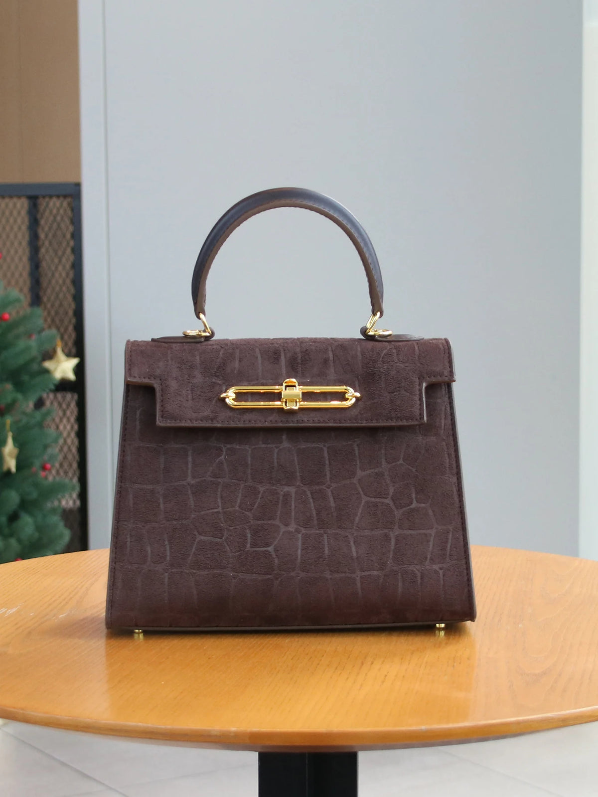 A dark brown crocodile pattern tote bag with a leather handle and gold hardware sitting on a wooden table.