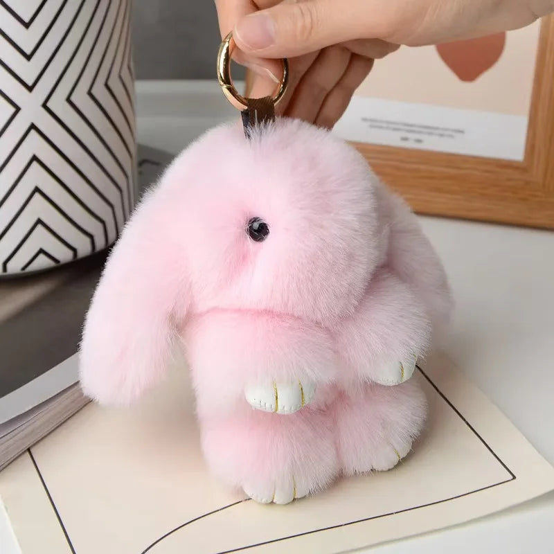 A pink fluffy rabbit plush charm attached to a gold keyring, resting on a light-colored paper.