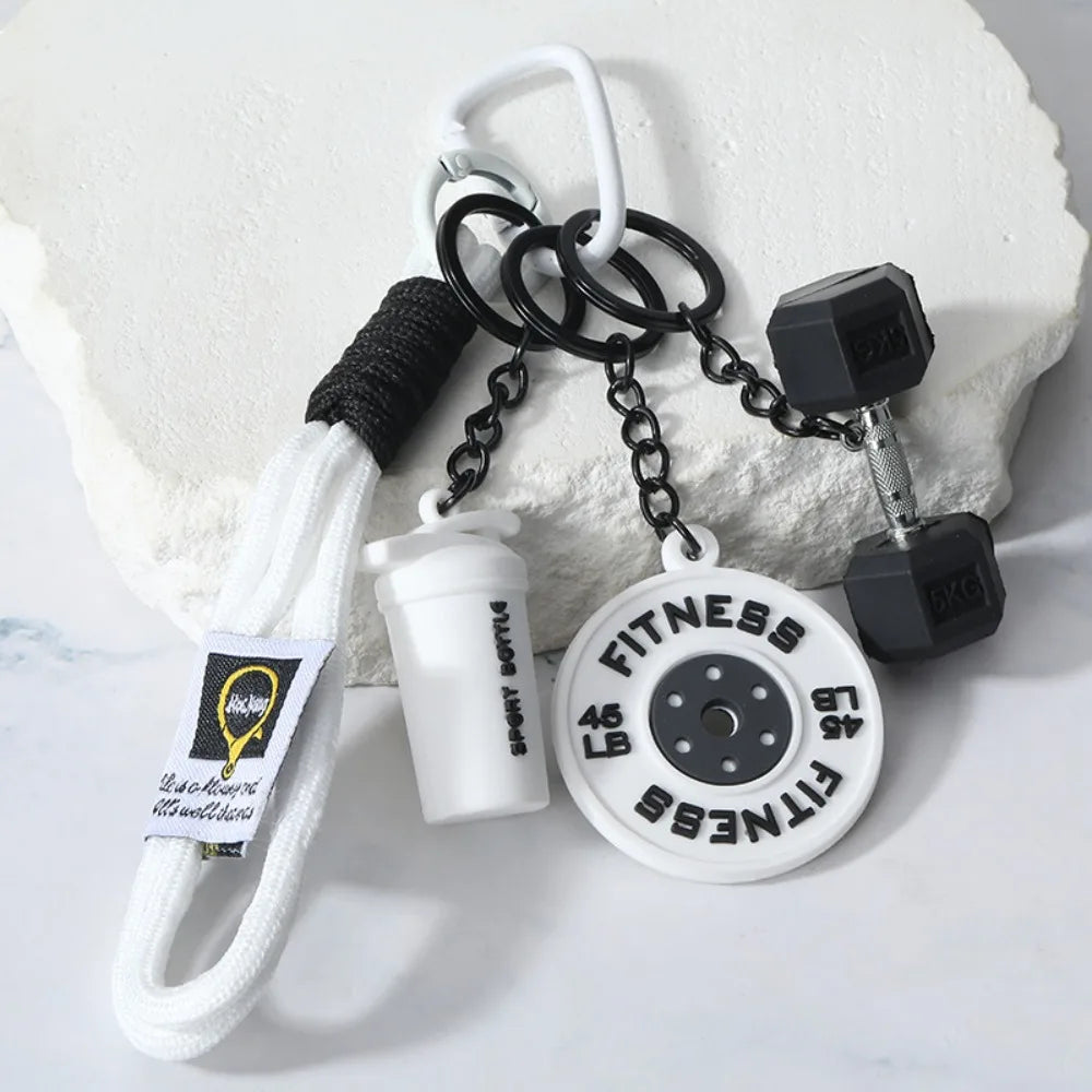 A white fitness dumbbell charm keychain with a black grip and chain is displayed on a white surface.