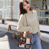Une femme en pull beige et jeans tient un sac à bandoulière en cuir marron devant une fenêtre en verre.