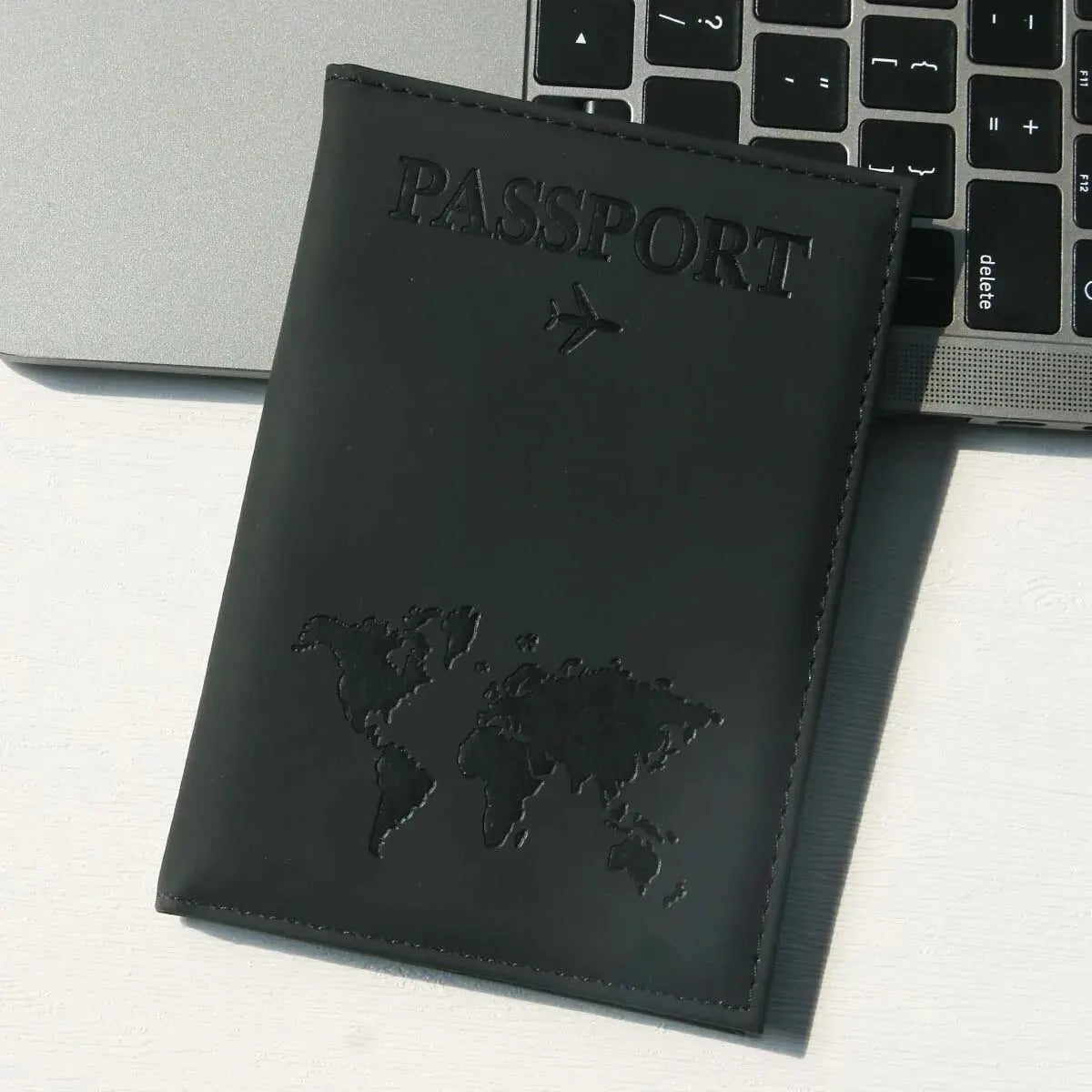 A black leather passport holder with the word PASSPORT and a world map design resting on a white surface next to a laptop.