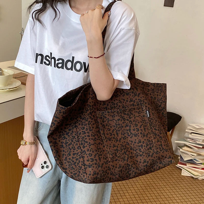 A person holds a brown leopard print canvas tote bag against a white t-shirt.