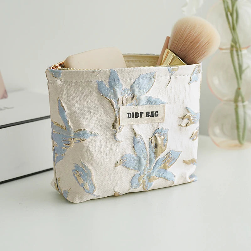 A beige canvas makeup bag with gold and blue floral embroidery is shown next to a brush.