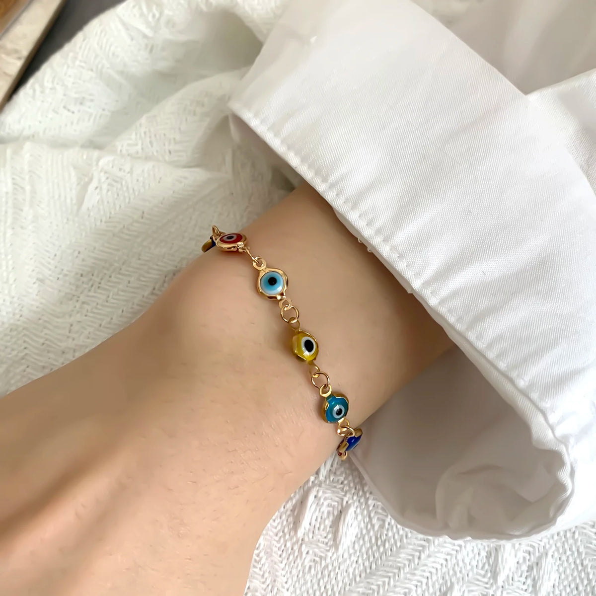 A close-up of a hand wearing a gold charm bracelet featuring blue evil eye beads against a white fabric background.