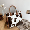 A brown leather tote bag with a white leopard print faux fur exterior resting on a wooden surface.