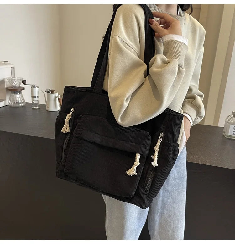A person holds a black corduroy shoulder bag with gold-colored charms against a light background.