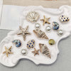 A collection of gold and silver conch shell and starfish jewelry charms displayed on a white ceramic dish.