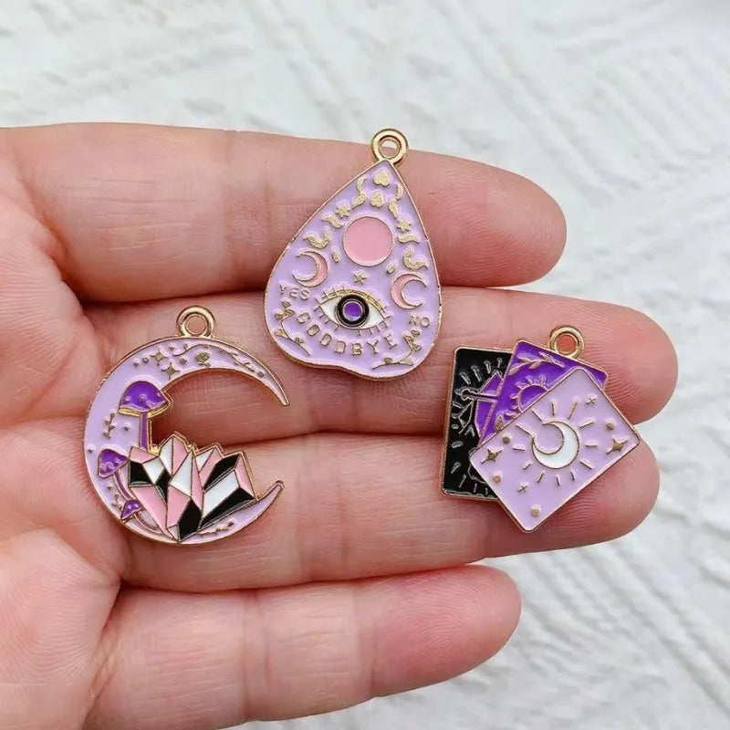 A hand holds three celestial tarot charms featuring pink and purple stones, including a teardrop pendant and a crescent moon 