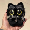 Colgante de peluche de gato negro con grandes ojos amarillos y una púa de guitarra plateada.
