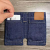A close-up of a dark blue denim mini wallet and card holder with a leather patch on the top right pocket.