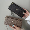 A hand holds a dark brown patterned wallet with gold zipper and leather strap against a gray background.