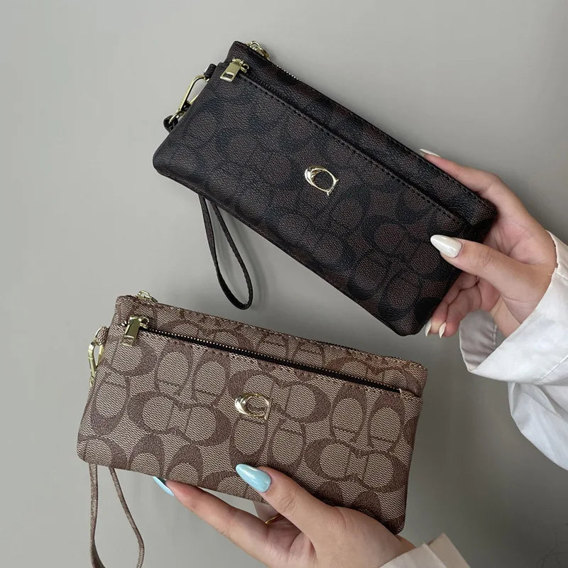 A hand holds a dark brown patterned wallet with gold zipper and leather strap against a gray background.