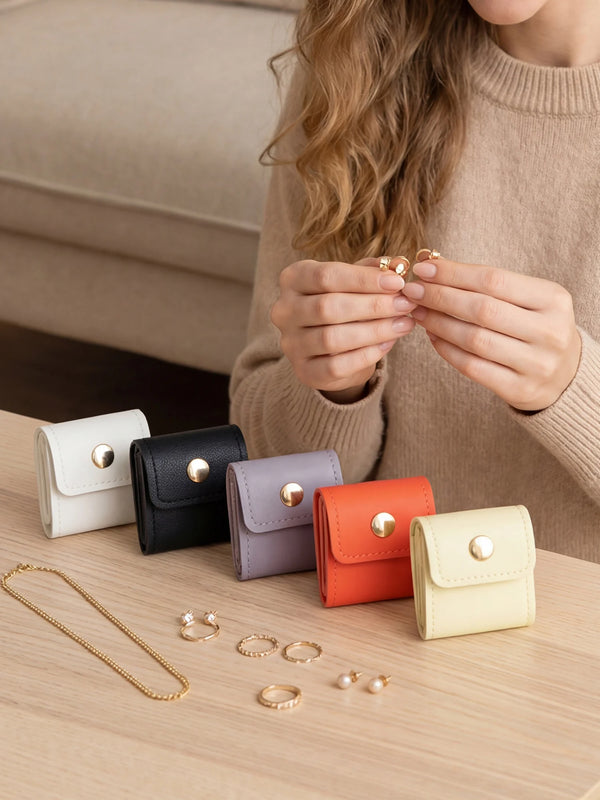 A person holds a gold ring and several small gold rings over a light wooden surface next to folded mini jewelry storage pouch