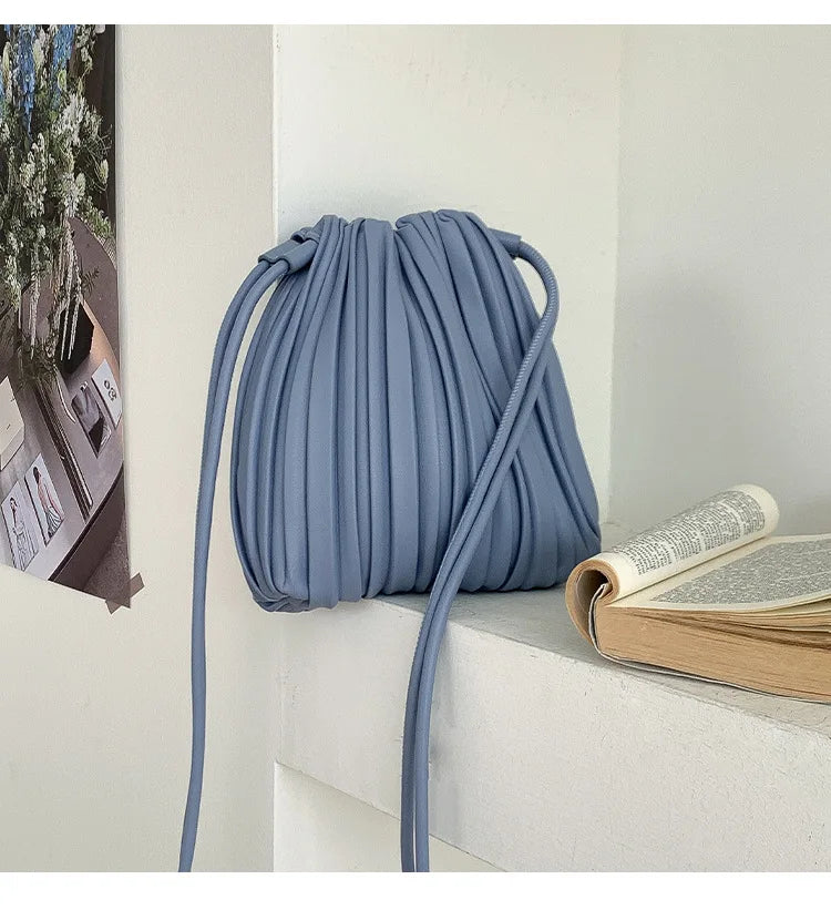 A light blue PU leather pleated crossbody bag hanging on a white shelf next to a stack of books.