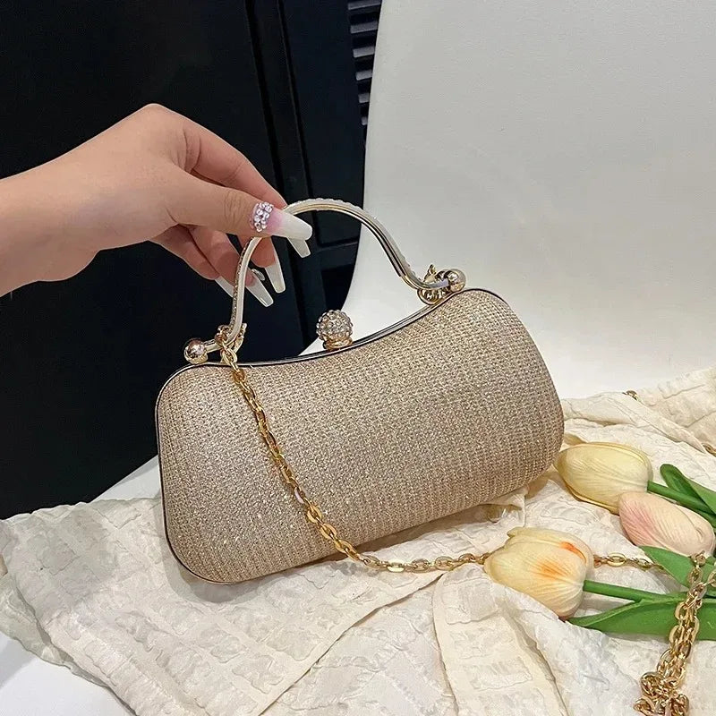 A hand holds a beige glitter crossbody evening clutch bag with gold chain details on a white textured surface.
