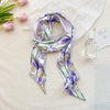 Écharpe florale de soie violette et verte drapée sur un tissu blanc à côté d'une bouteille de parfum.