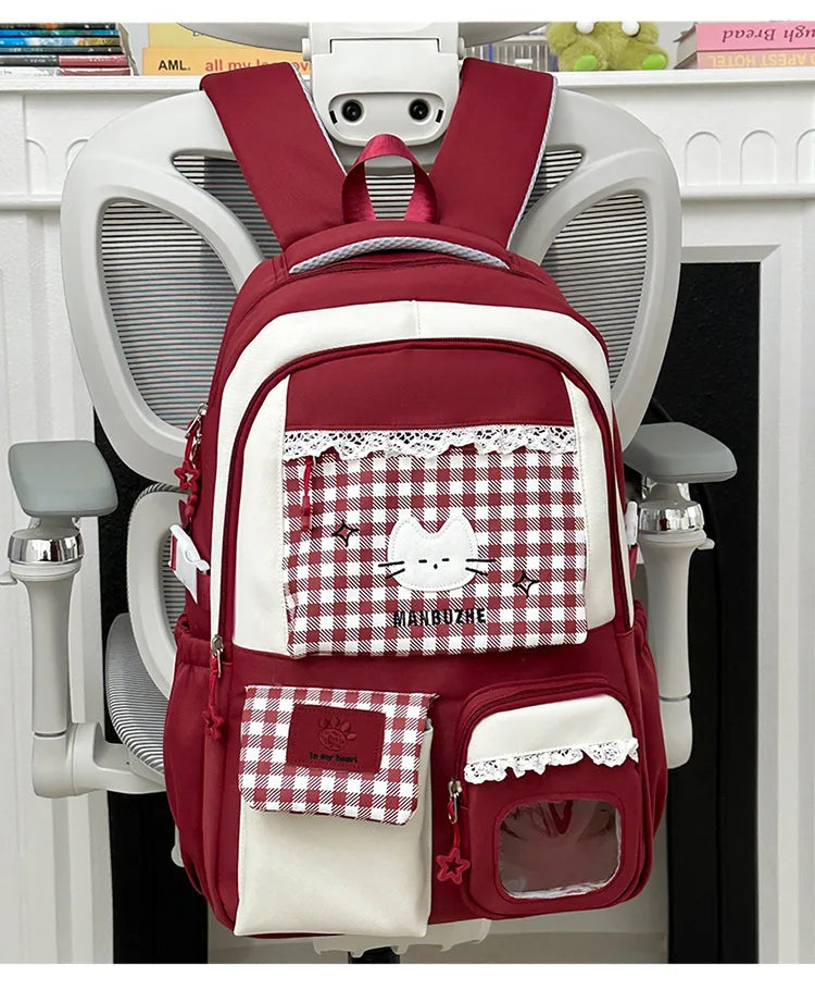 A maroon and white plaid backpack with a cat design and laptop compartment hanging on a white door.