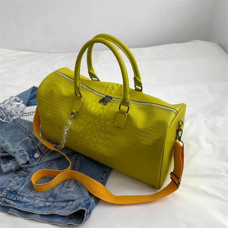 A lime green crocodile grain PU leather travel duffle bag with tan leather straps resting on a white bed next to denim jeans.