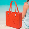 An orange portable EVA beach tote bag is shown resting on light sand with a blurred background of blue water.