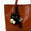 A black cat plush keychain charm with gold eyes attached to a brown leather handbag.