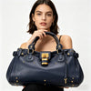 A woman holds a navy blue vegan leather tote bag with gold hardware against a white background.
