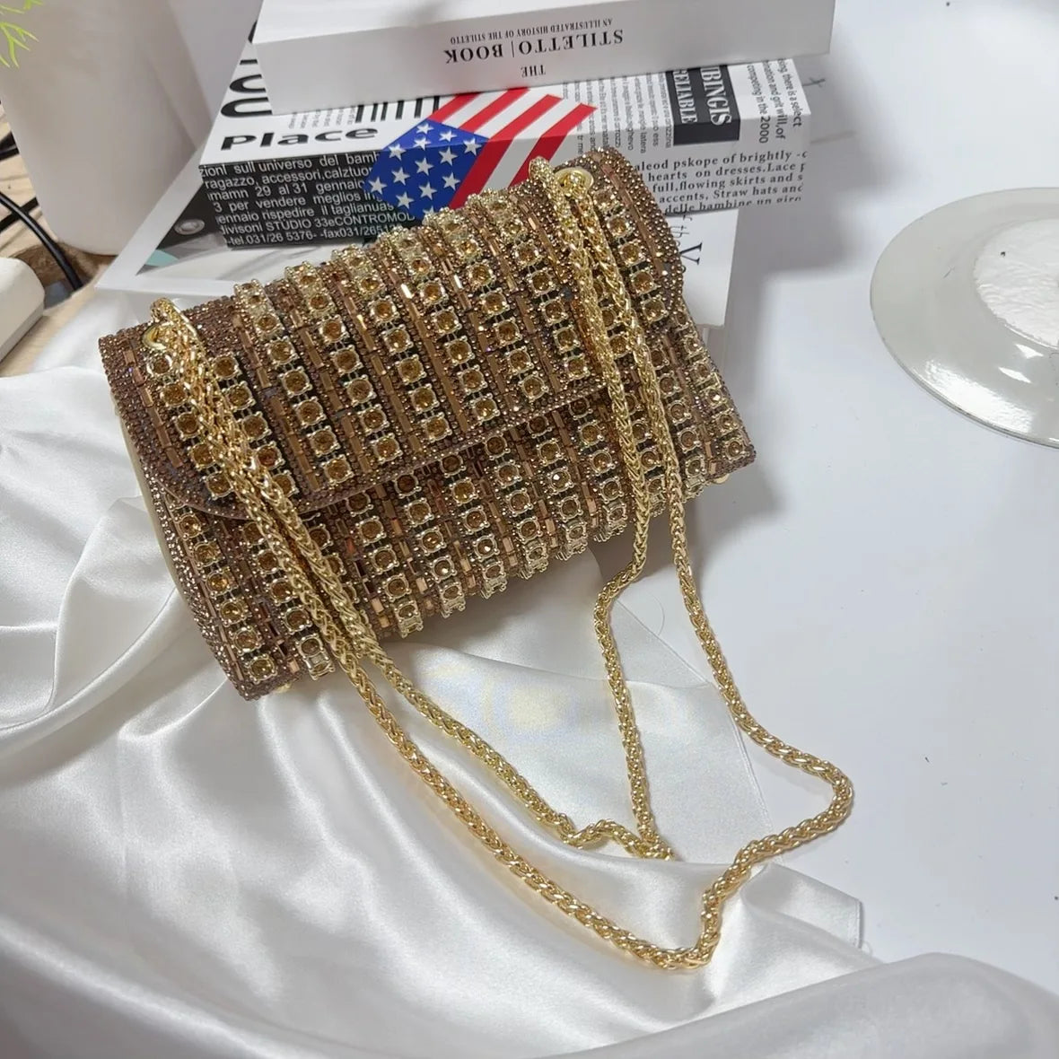 A gold rhinestone evening clutch bag with a textured pattern is displayed on white satin fabric.