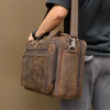 A brown genuine leather briefcase with dark stitching is carried by a person wearing a black shirt and gray pants.
