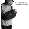 A person wearing a gray jacket is demonstrating a black sling bag designed for phone and wallet access.