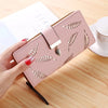 Clutch cartera de cuero sintético rosa con estampado de hojas grabadas, sostenida por una mano sobre una superficie de madera