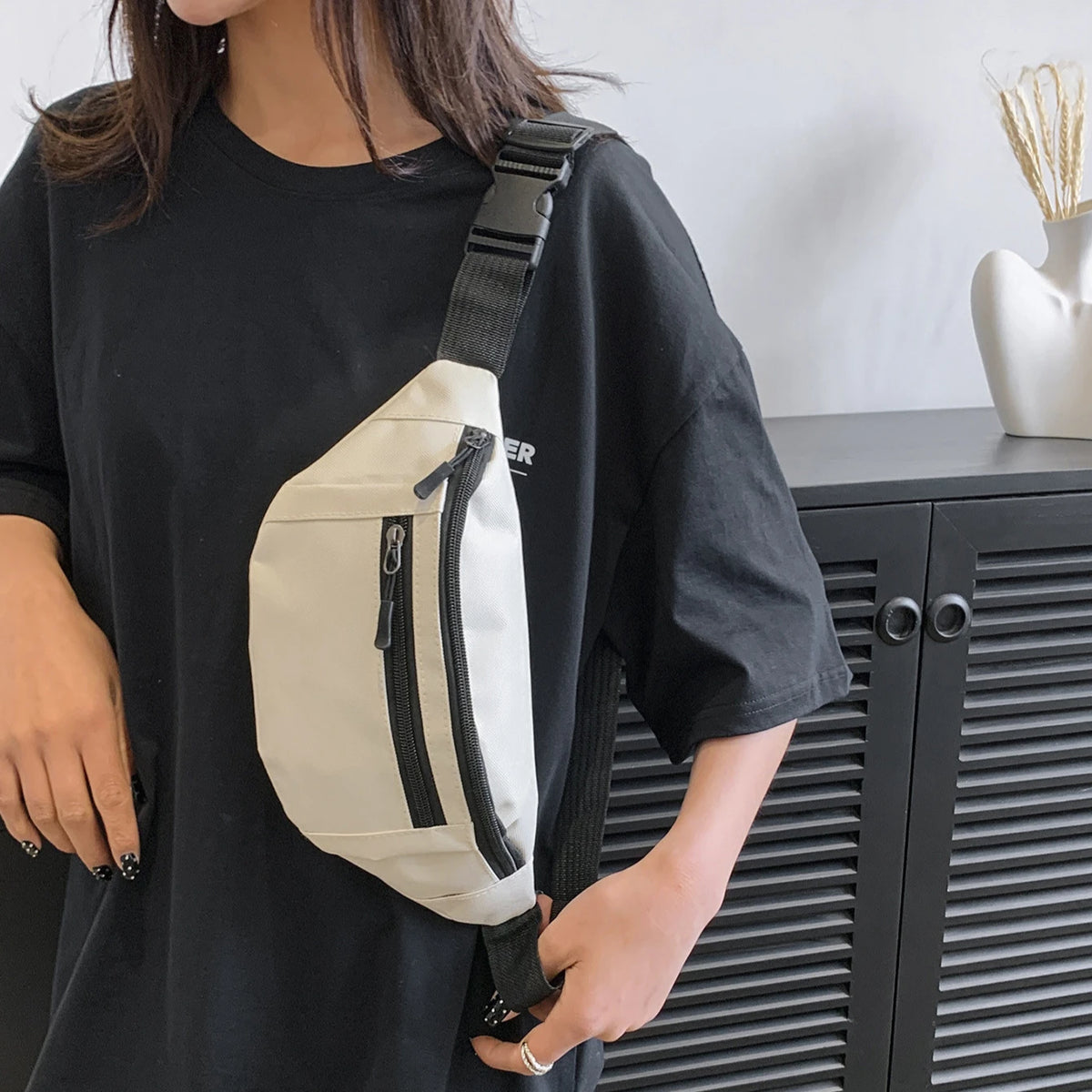 A person is holding a beige nylon waist bag with black zippers against a black t-shirt.