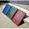 Three genuine leather passport holders in blue, green, and red stacked on a light wooden surface.
