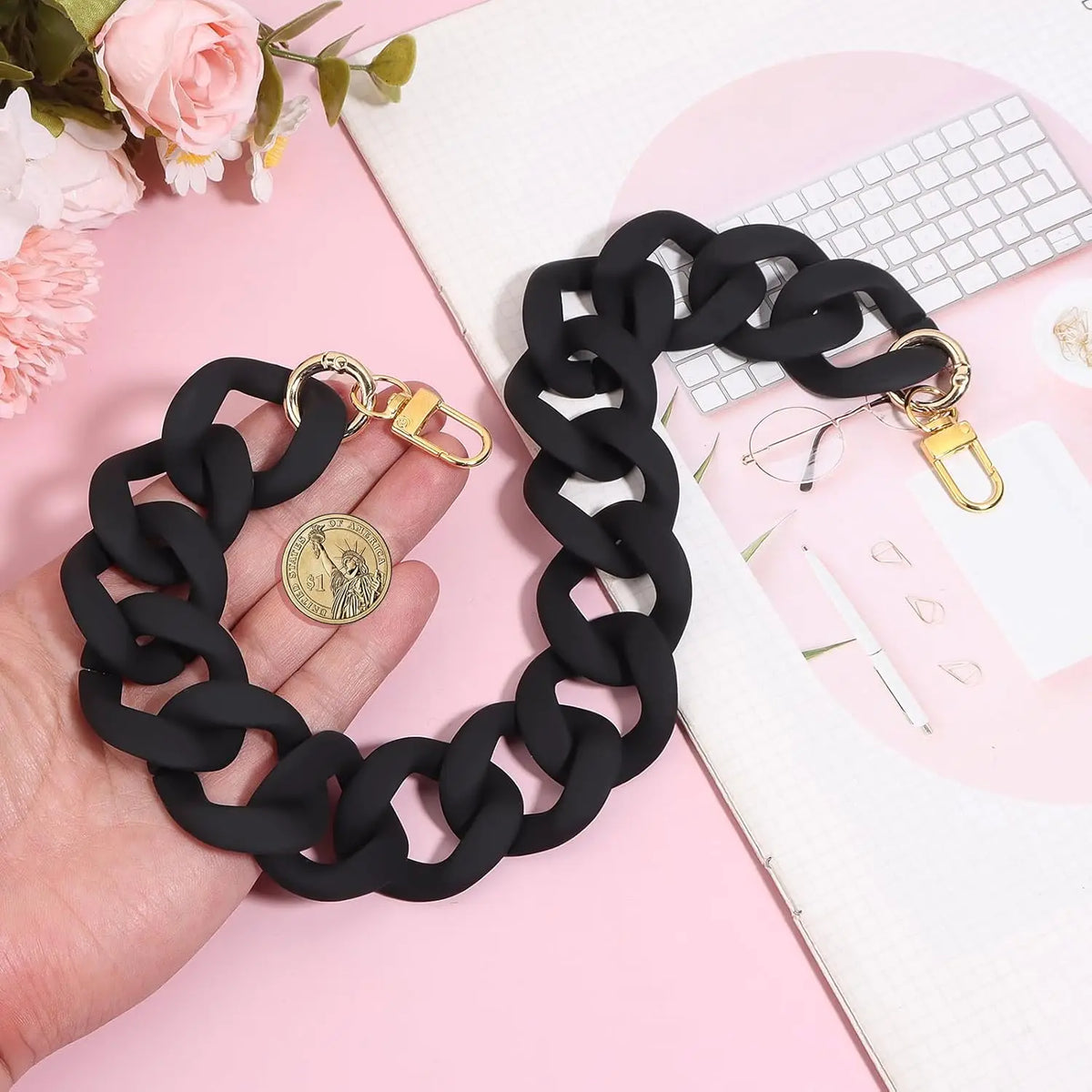 Black acrylic chain strap with gold clasps displayed on a pink background.
