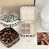 A white leopard print jewelry storage box is shown open, next to a folded dark brown leopard print pouch and gold rings.