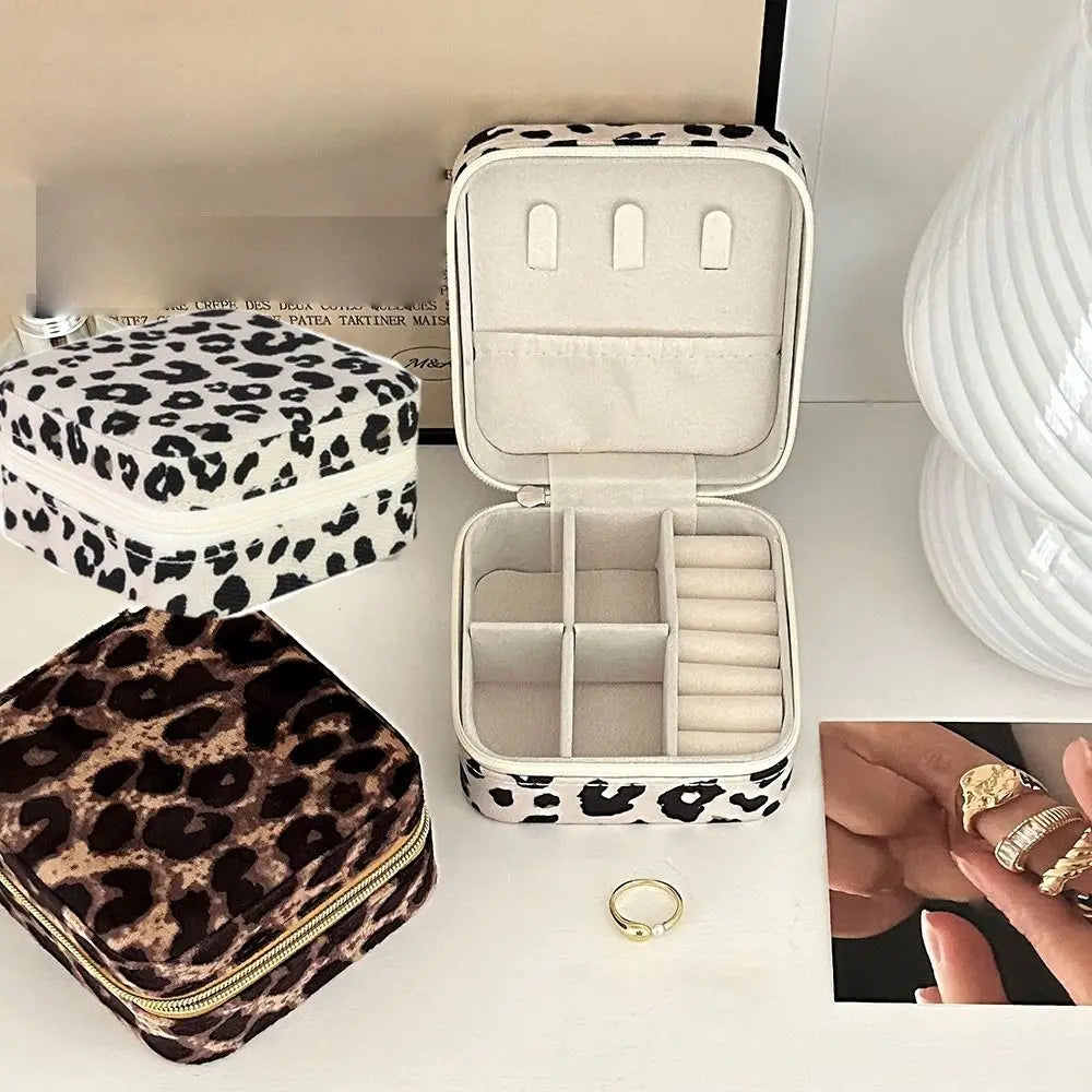 A white leopard print jewelry storage box is shown open, next to a folded dark brown leopard print pouch and gold rings.