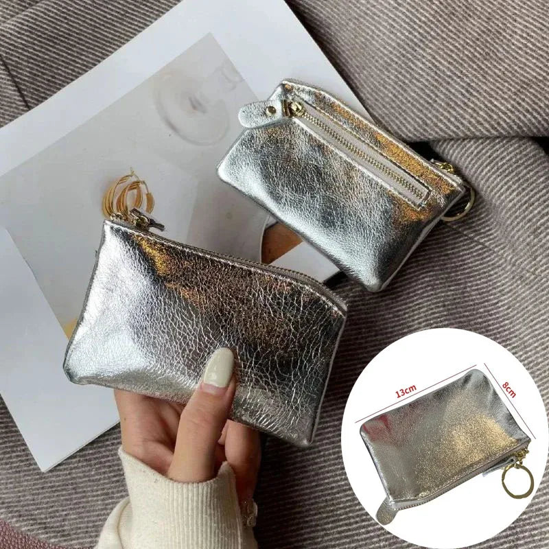 A silver metallic leather compact coin purse and key holder is shown being held against a gray fabric background.