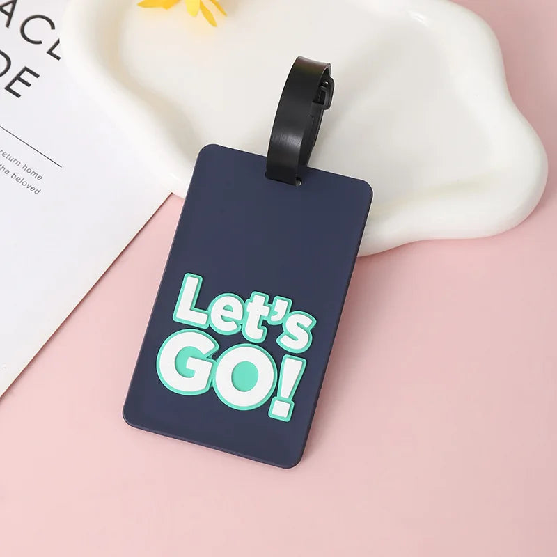 A dark blue silicone luggage tag with the text Let's GO! is displayed on a pink background.