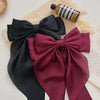 A close-up of a dark maroon silk bow hair clip and a black silk ribbon, resting on a light beige textured fabric.