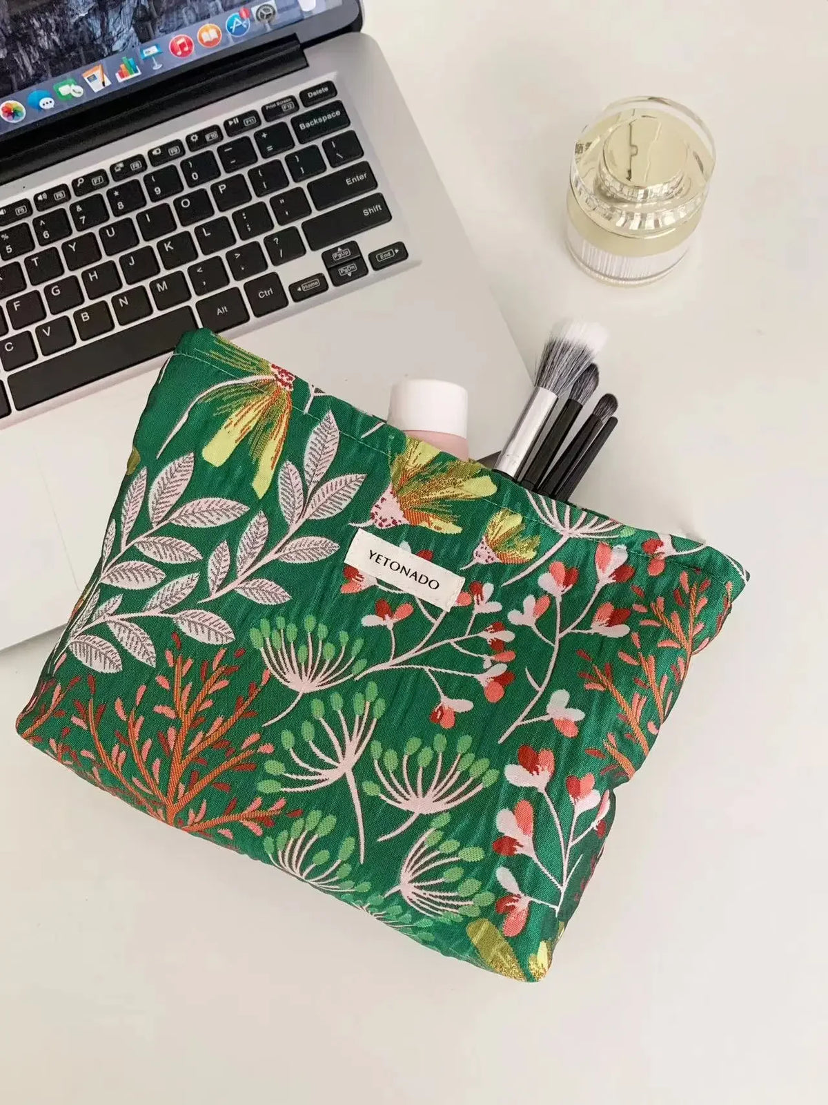 A green cosmetic pouch with floral embroidery rests on a white desk next to a laptop and makeup brushes.