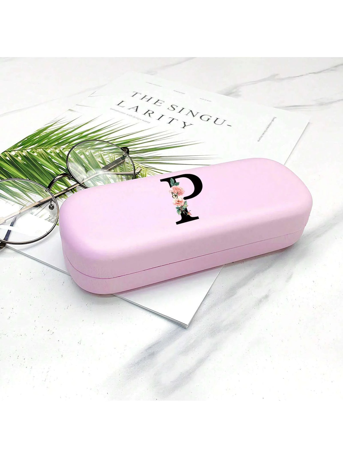 A pink hard shell eyeglass case with a letter charm rests on a white marble surface next to a pair of eyeglasses.