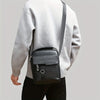 Un homme portant un pull gris clair et un pantalon noir, tenant un sac à bandoulière en PU noir.