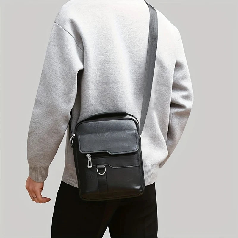 Un homme portant un pull gris clair et un pantalon noir, tenant un sac à bandoulière en PU noir.